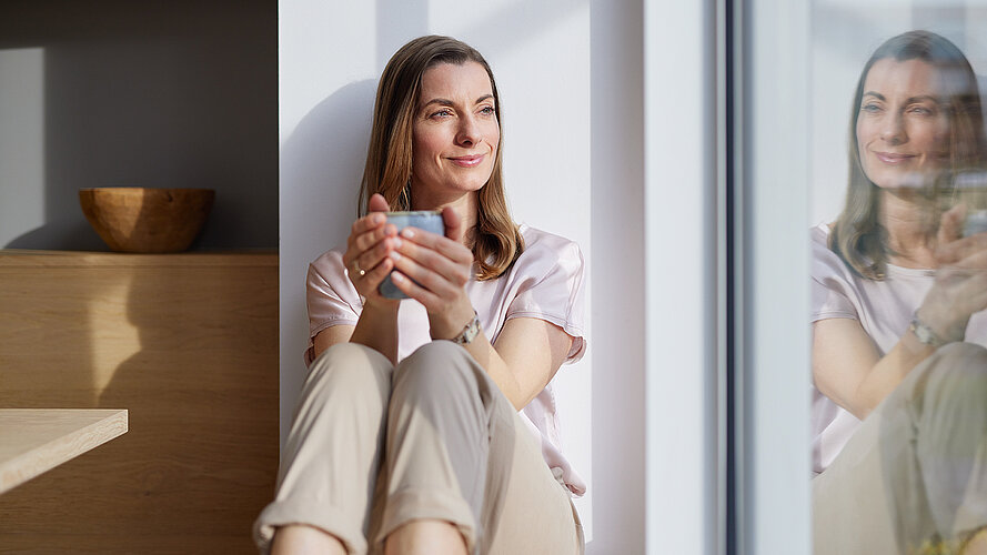 Frau sitzt auf der Fensterbank mit einer Tasse und schaut aus dem Fenster in welchem sie sich spiegelt.